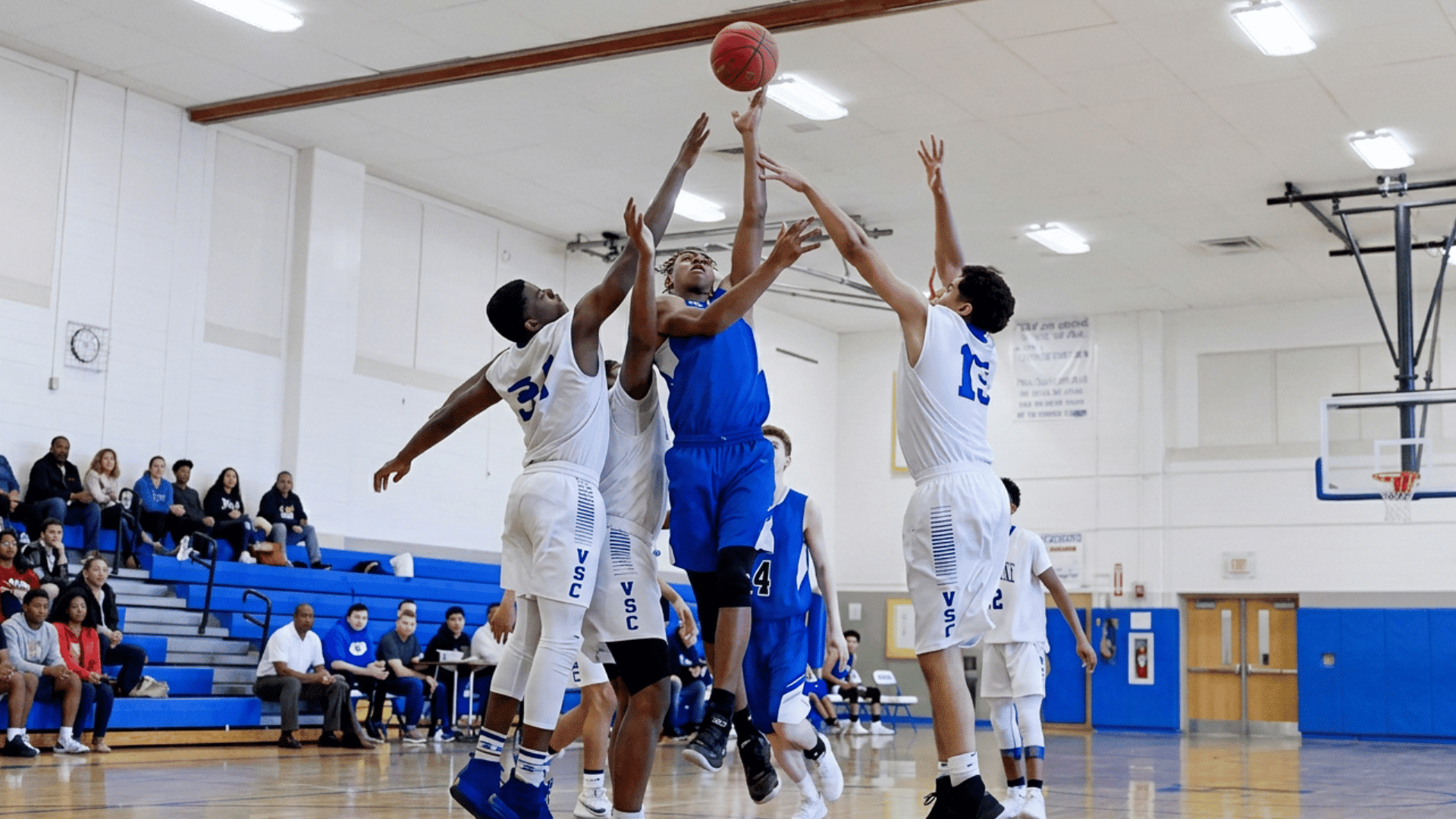 basketball player in blue jersey jumps for a shot between three defenders in white uniforms during a game in a school gym with spectators (1)
