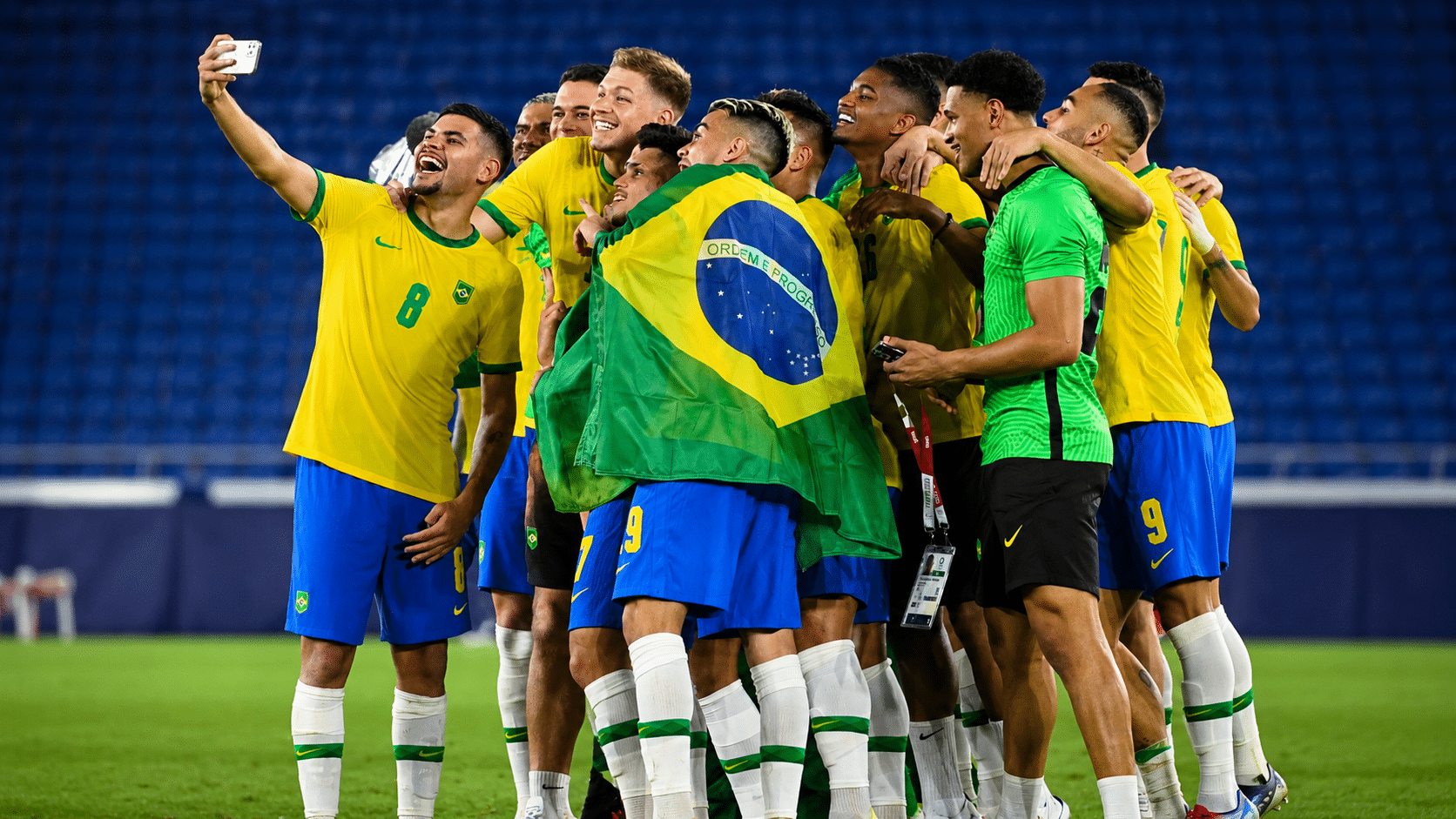 brazil football players huddle together on the pitch smiling while taking a celebratory selfie with a brazilian flag draped over one player