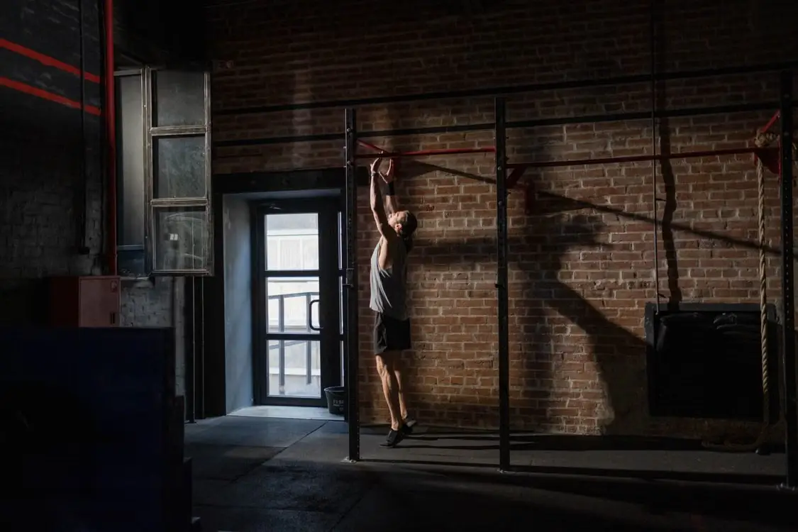 Man reaching for pull-up bar in dimly lit gym with brick walls