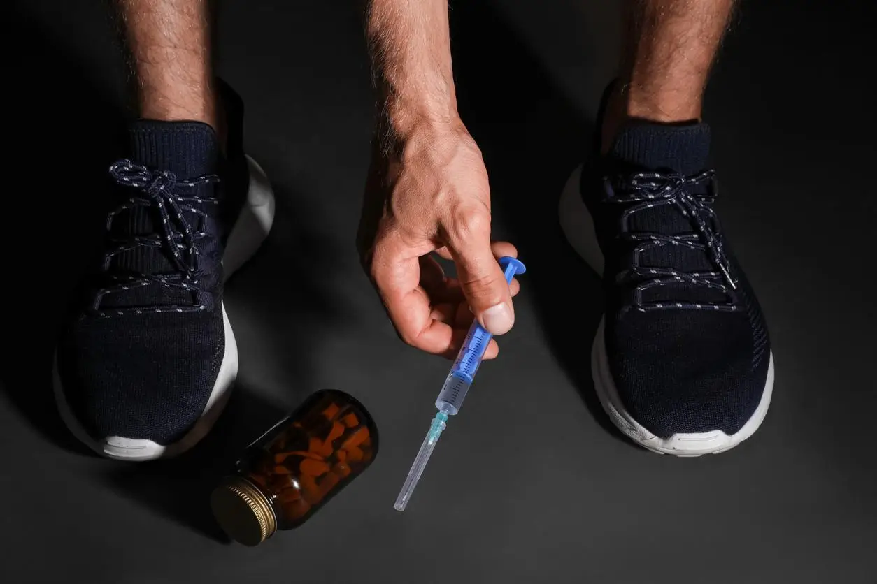 Hand holding syringe next to pill bottle and running shoes on dark surface