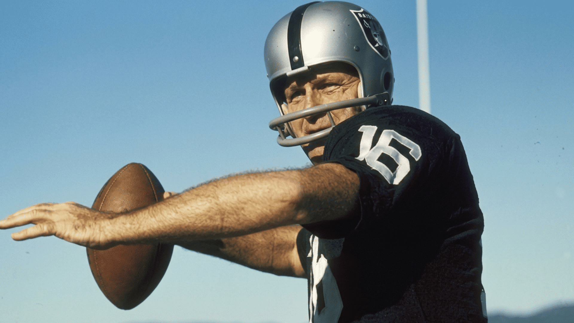 football player in a black jersey and silver helmet prepares to throw a football with one arm extended against a clear sky