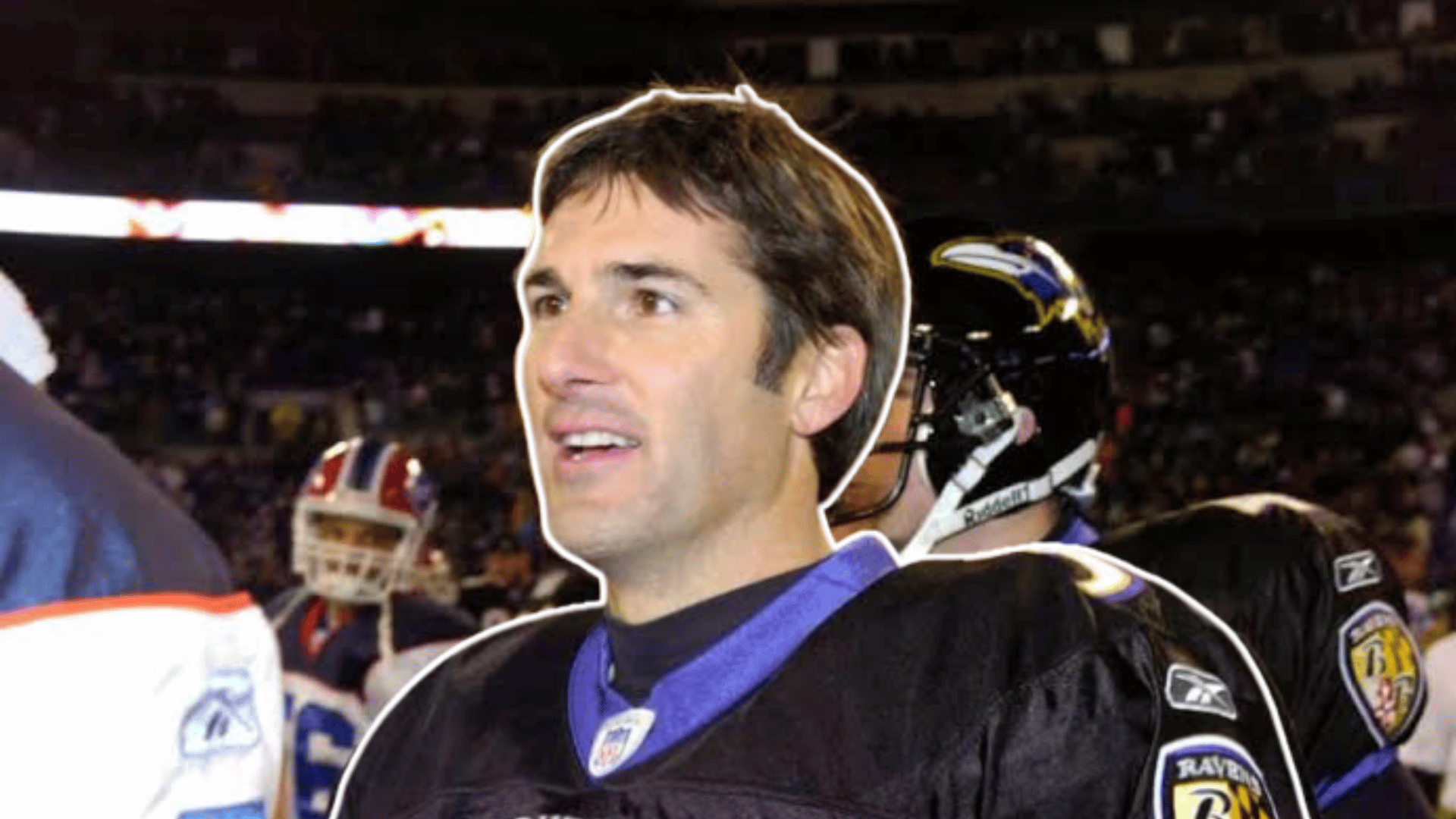 football player in a black jersey stands on the field smiling before the game with teammates and stadium crowd in the background