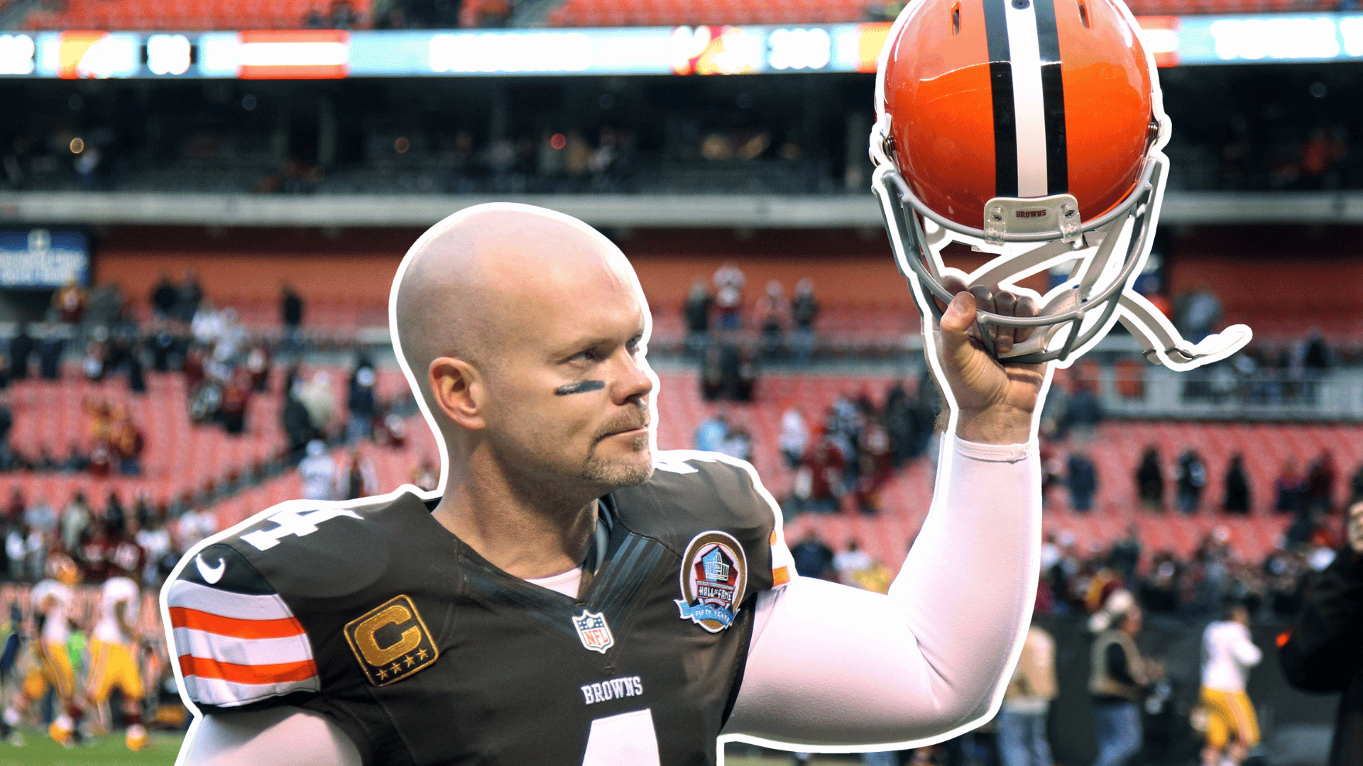 football player in a brown jersey raises an orange helmet after the game while standing on the field in a mostly empty stadium