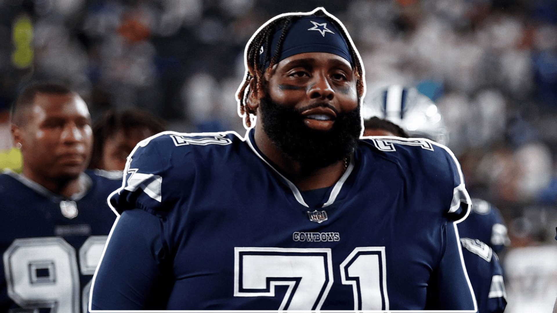 football player in a navy jersey and headband stands on the sideline smiling before the game with teammates and stadium crowd behind him
