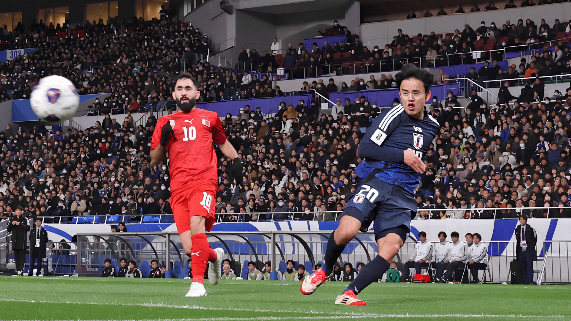 japan mens soccer player crosses the ball during a match as an opposing defender chases in front of a packed stadium crowd