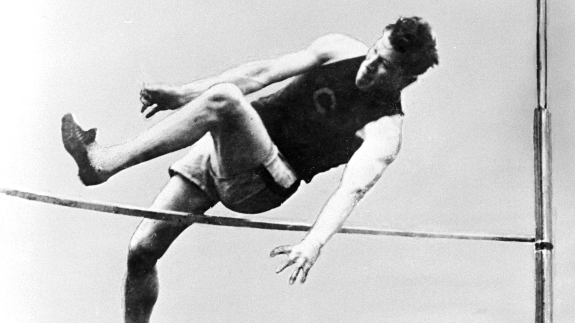 Jim Thorpe clearing the high jump bar during competition wearing a dark athletic uniform in a historic black and white photograph