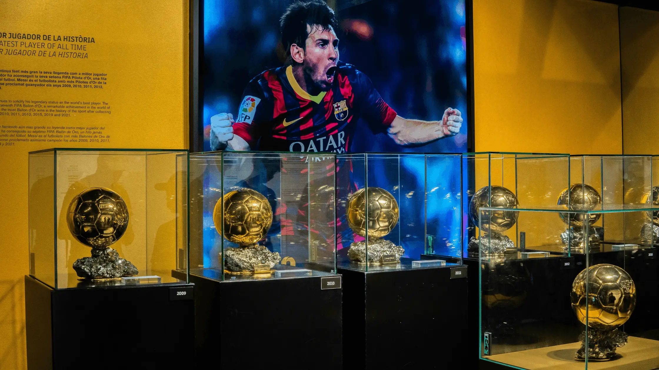 Glass display cases with multiple Ballon d'Or trophies and a large player poster on yellow wall