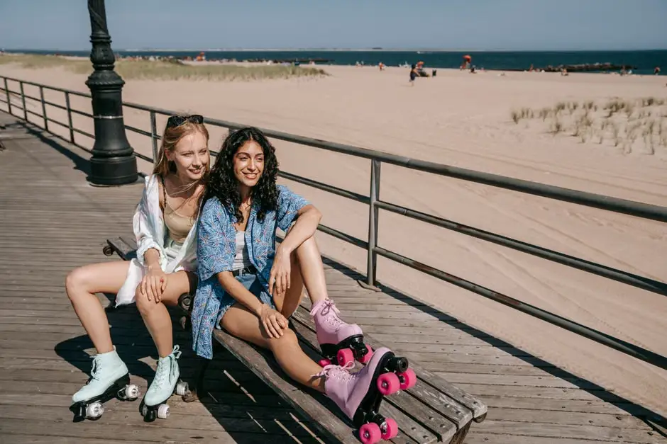 Two women sitting on boardwalk bench wearing roller skates by sandy beach