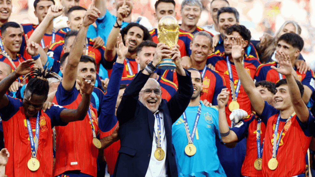 spain team members surround a smiling official holding the world cup trophy high while celebrating their championship victory