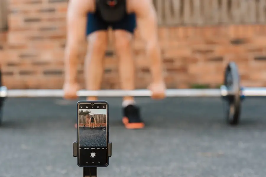 Man preparing to lift barbell outside with smartphone recording in foreground