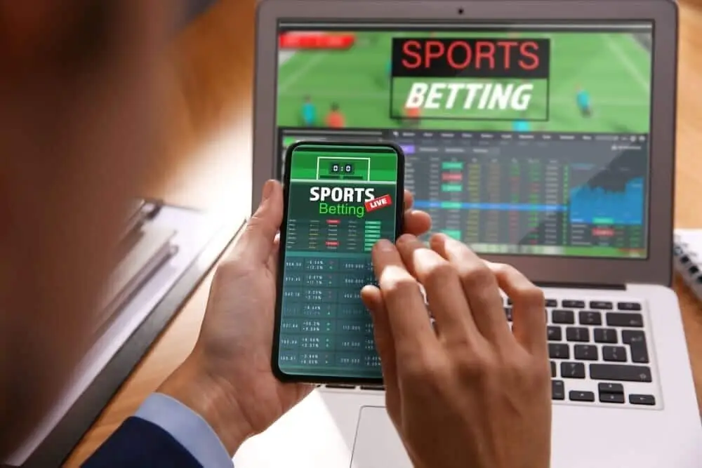 Smartphone displaying sports betting app in front of laptop with sports screen in office setting