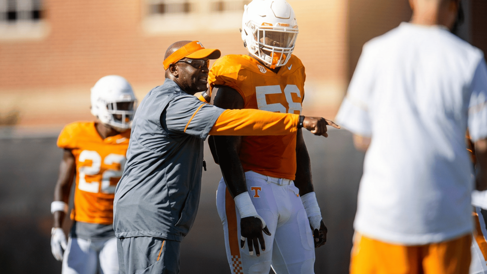 a football coach points across the practice field while speaking with a player in orange uniform during a daytime training session