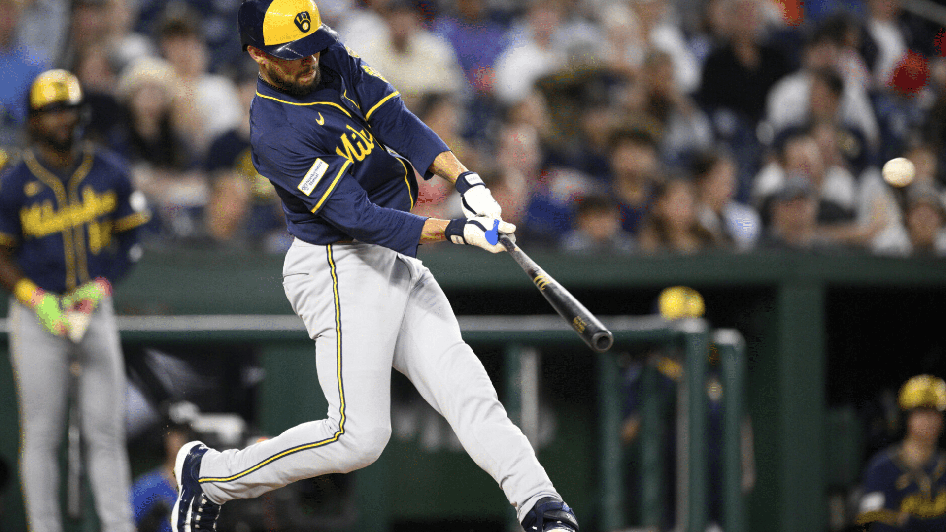 baseball player in a blue and yellow uniform swings the bat making contact with the ball during a game before a crowd