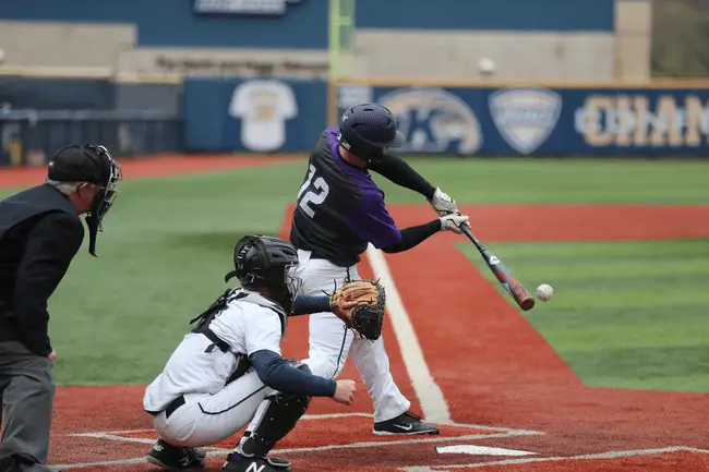 Baseball player in purple jersey swinging bat on outdoor field with catcher and umpire nearby