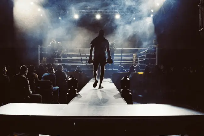Boxer walking towards ring under dramatic stage lights in smoky arena atmosphere