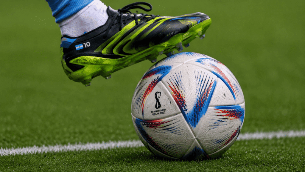 close up soccer scene showing a black and neon green football boot above a white ball with blue red patterns on a green field.