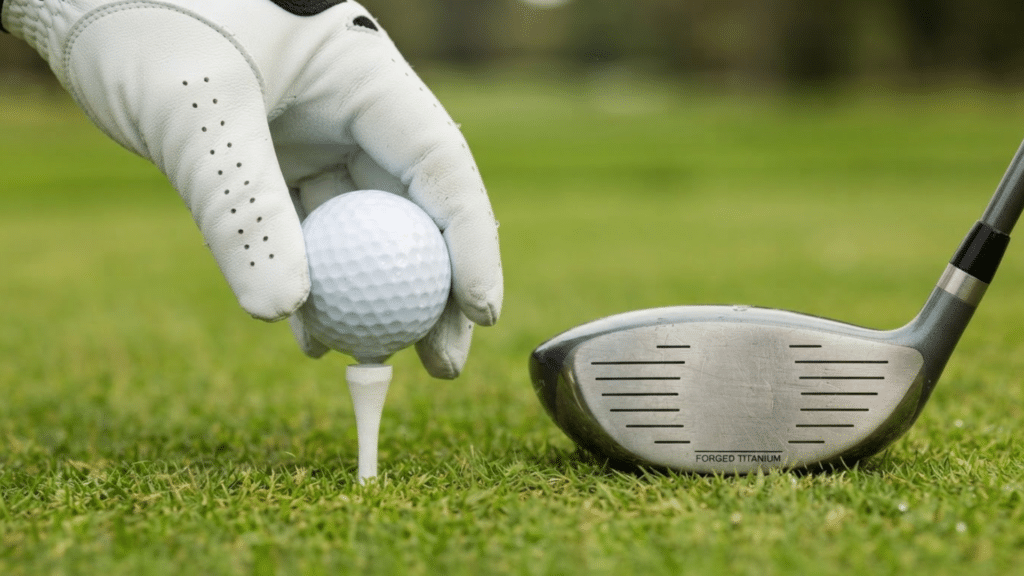 gloved hand placing a golf ball on a tee beside a driver on a well maintained golf course fairway