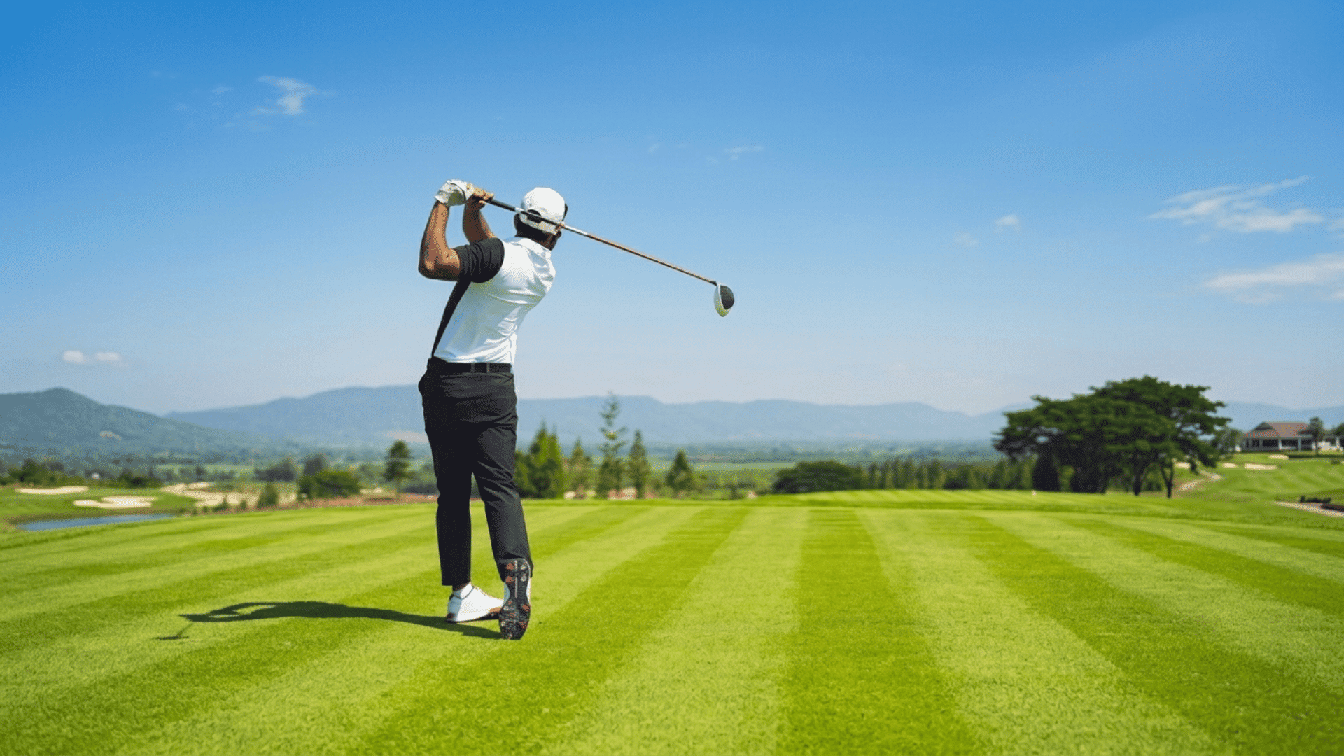 golfer swinging a driver on a well maintained fairway with mountains and clear blue sky in the distance