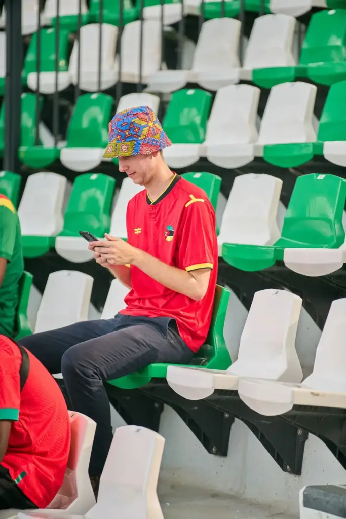 Young man in colorful hat using phone in stadium seating with green and white chairs