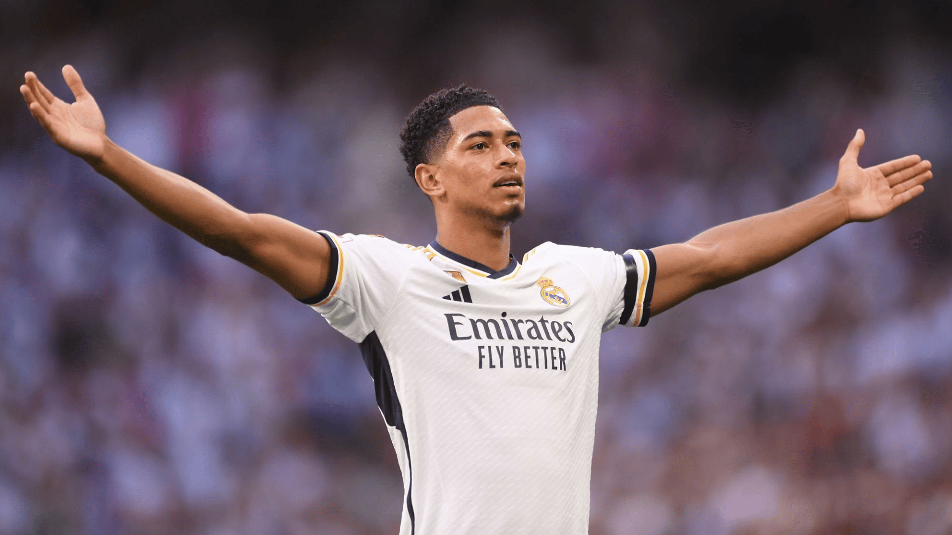 jude bellinghum wearing a white club jersey celebrating with arms spread wide on the field during a stadium match