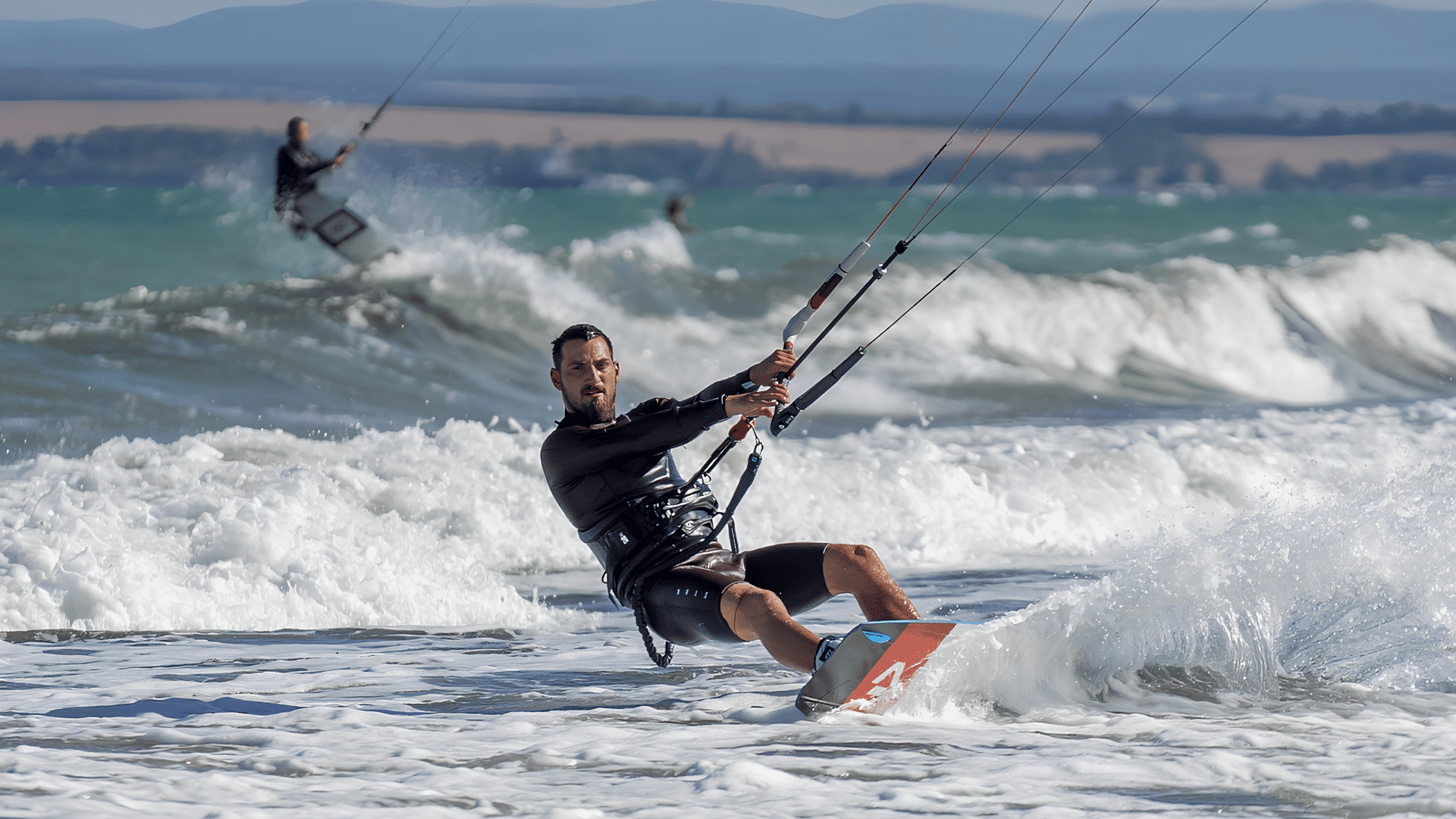 kitesurfer carving through ocean waves while holding the control bar on a windy day with other riders behind