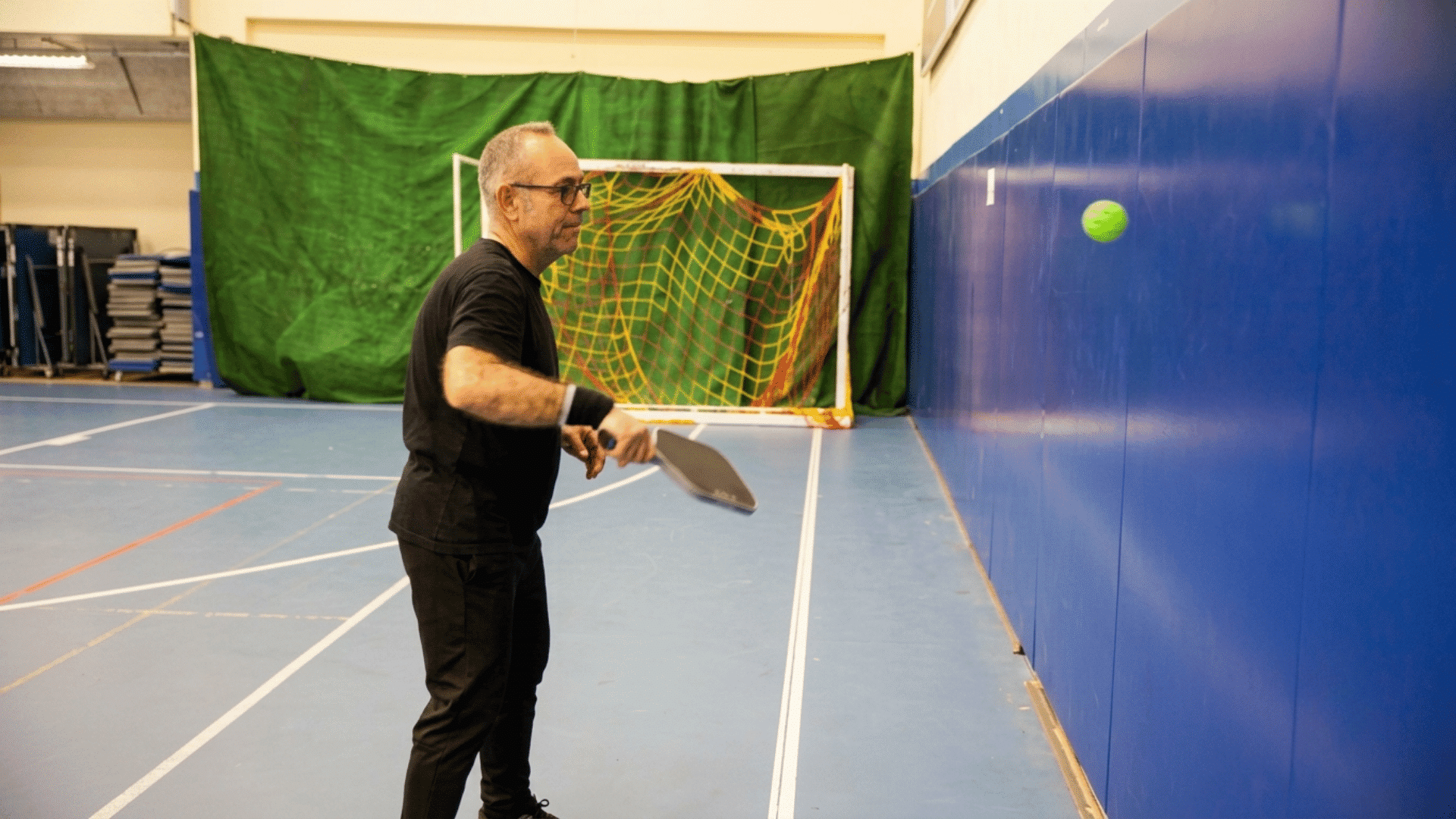 man practicing pickleball by hitting a ball against an indoor training wall with a sports net in the background