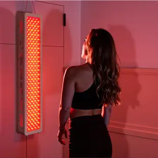 Woman in activewear standing near wall-mounted infrared light panel in dimly lit room