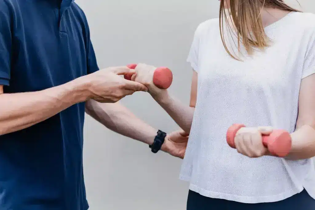 Person holding pink dumbbells with assistance from a trainer in an outdoor setting
