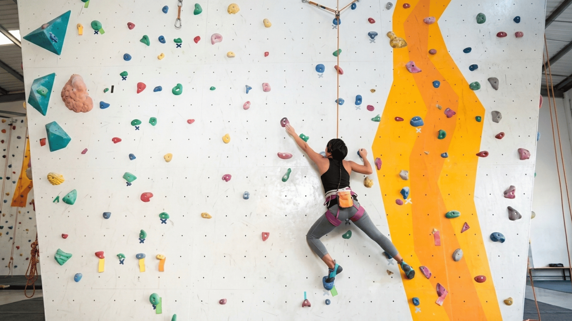 person climbing an indoor rock wall while reaching for colorful holds and wearing a harness in a climbing gym