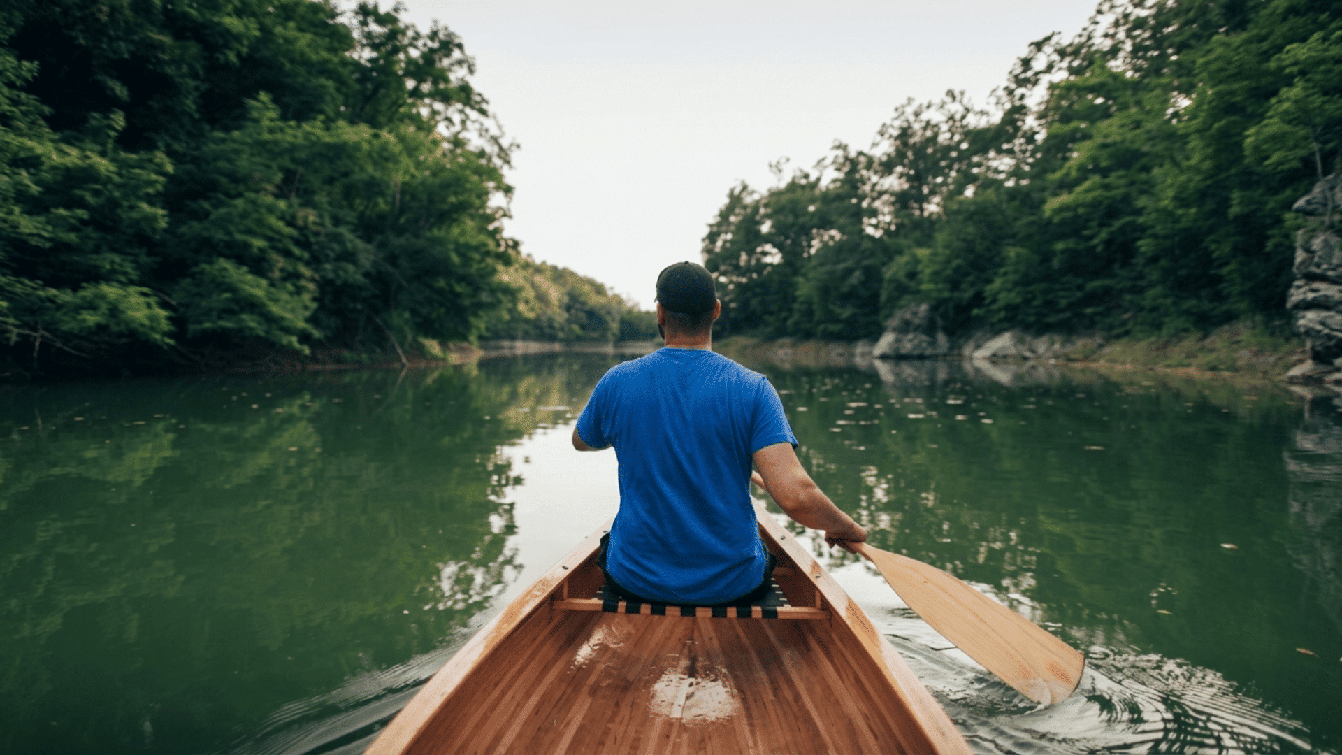 person paddling a wooden canoe along a calm river surrounded by dense green trees viewed from behind on a peaceful day
