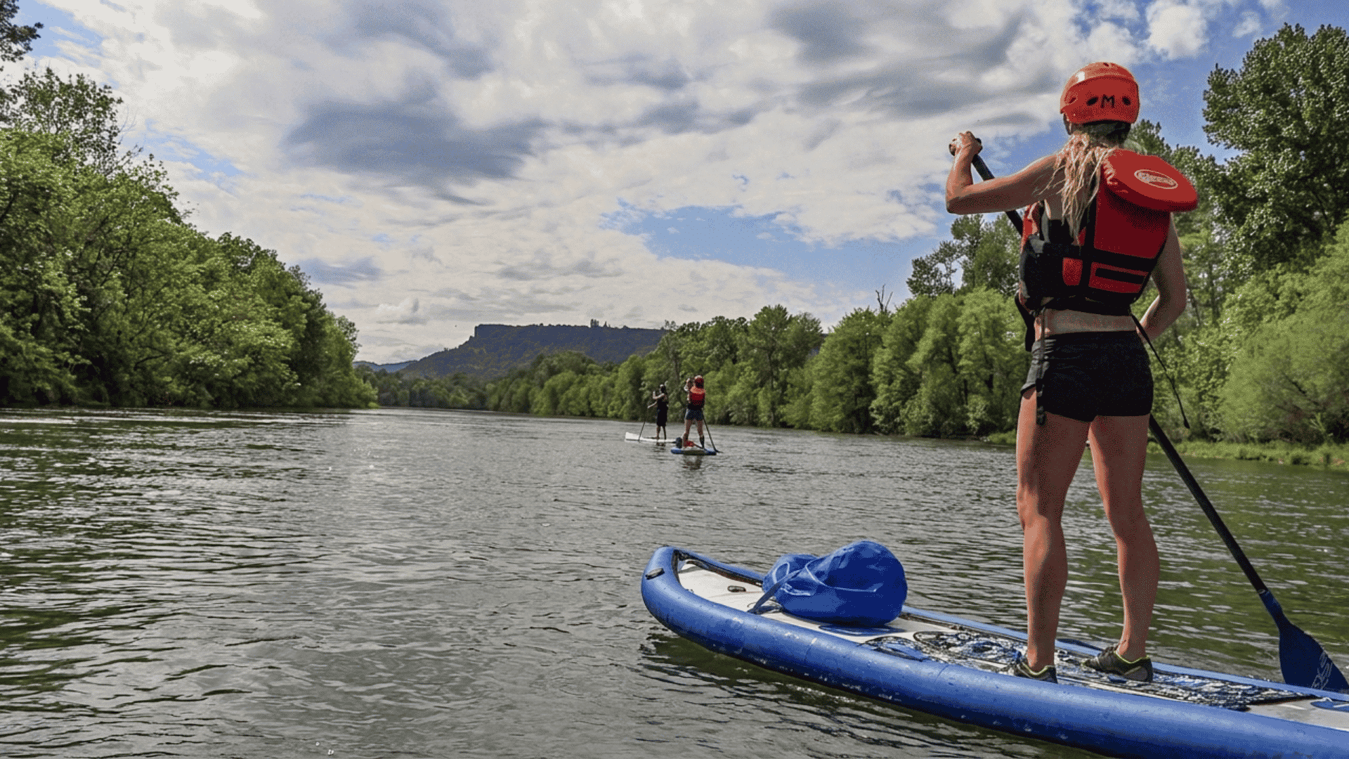 Person paddleboarding on a calm river while wearing a helmet and life jacket