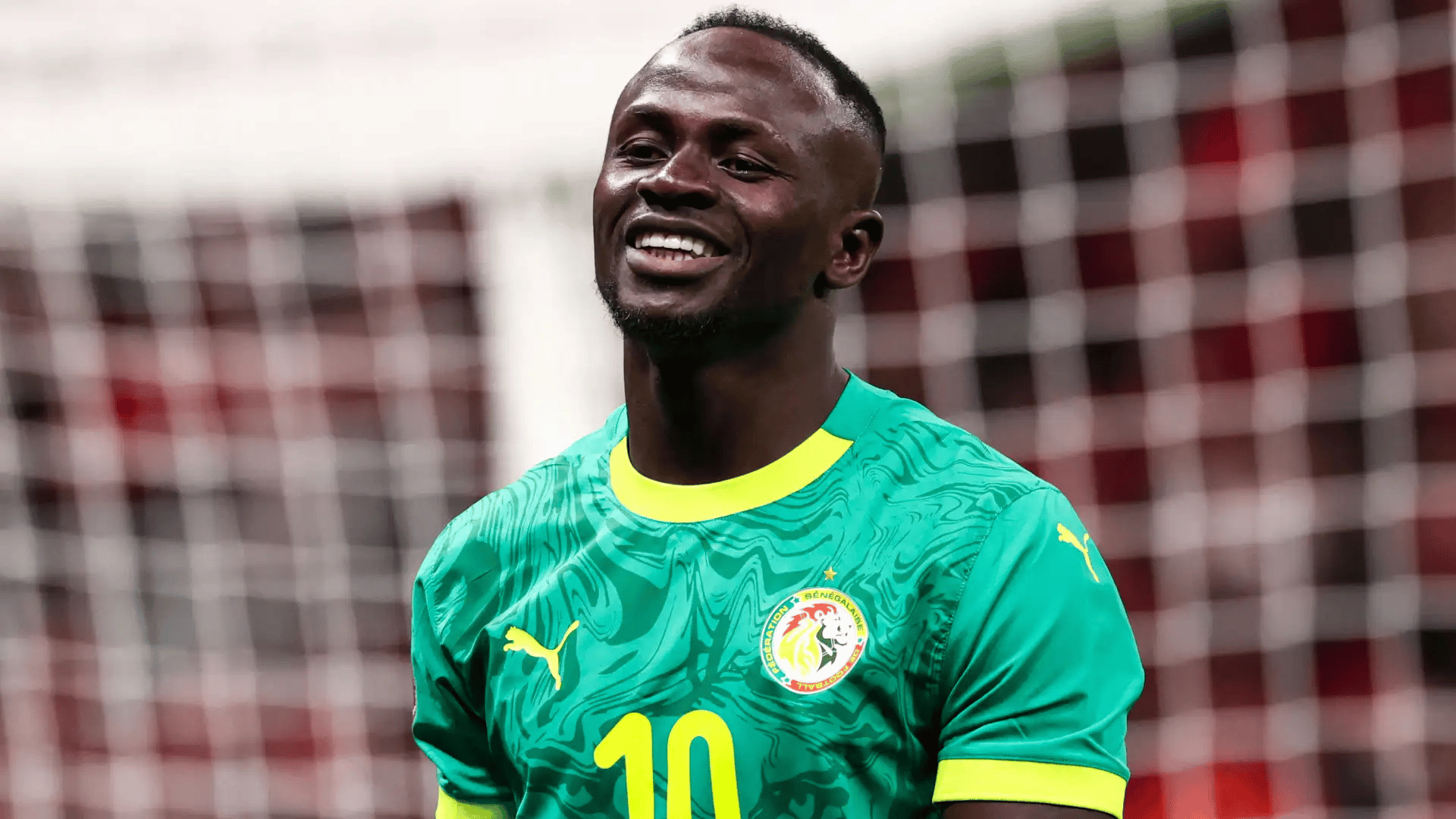 sadio mane wearing a green national team jersey smiling in front of a goal net during a stadium match