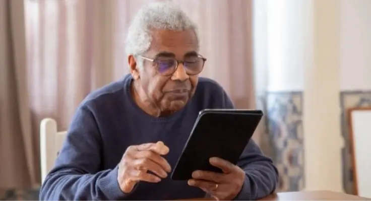 Older man in glasses using a tablet device at a wooden table indoors