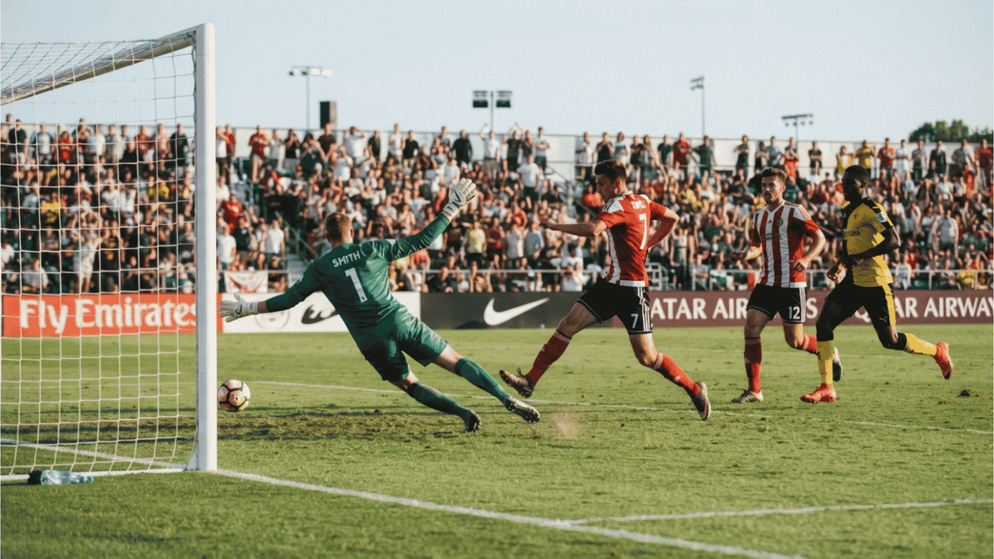 soccer player scores a goal past the diving goalkeeper while teammates and spectators watch the match unfold