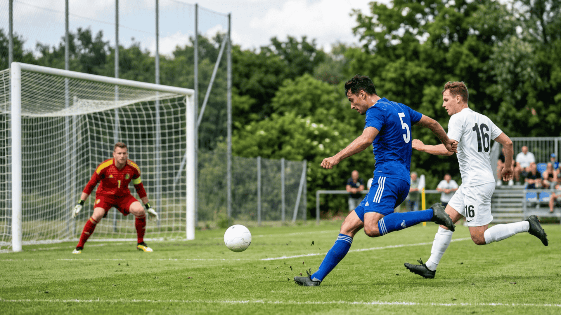 Soccer player in blue shoots on goal as a defender chases and the goalkeeper prepares to make a save.