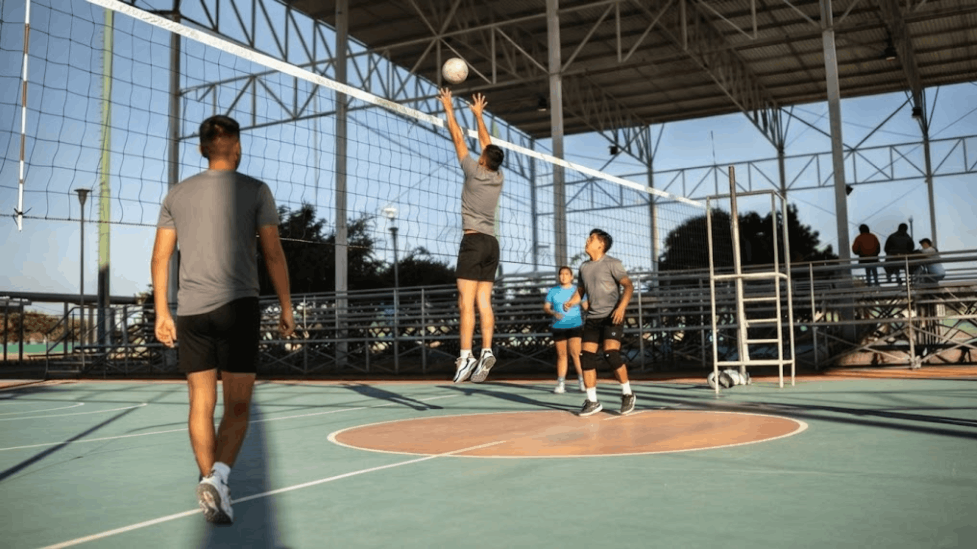 Volleyball players practicing at an indoor court with one player jumping to block the ball at the net during a training session under a covered arena
