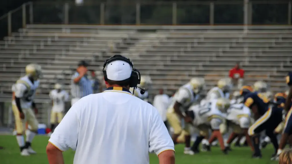 Football coach in white shirt observing players on field during daytime game