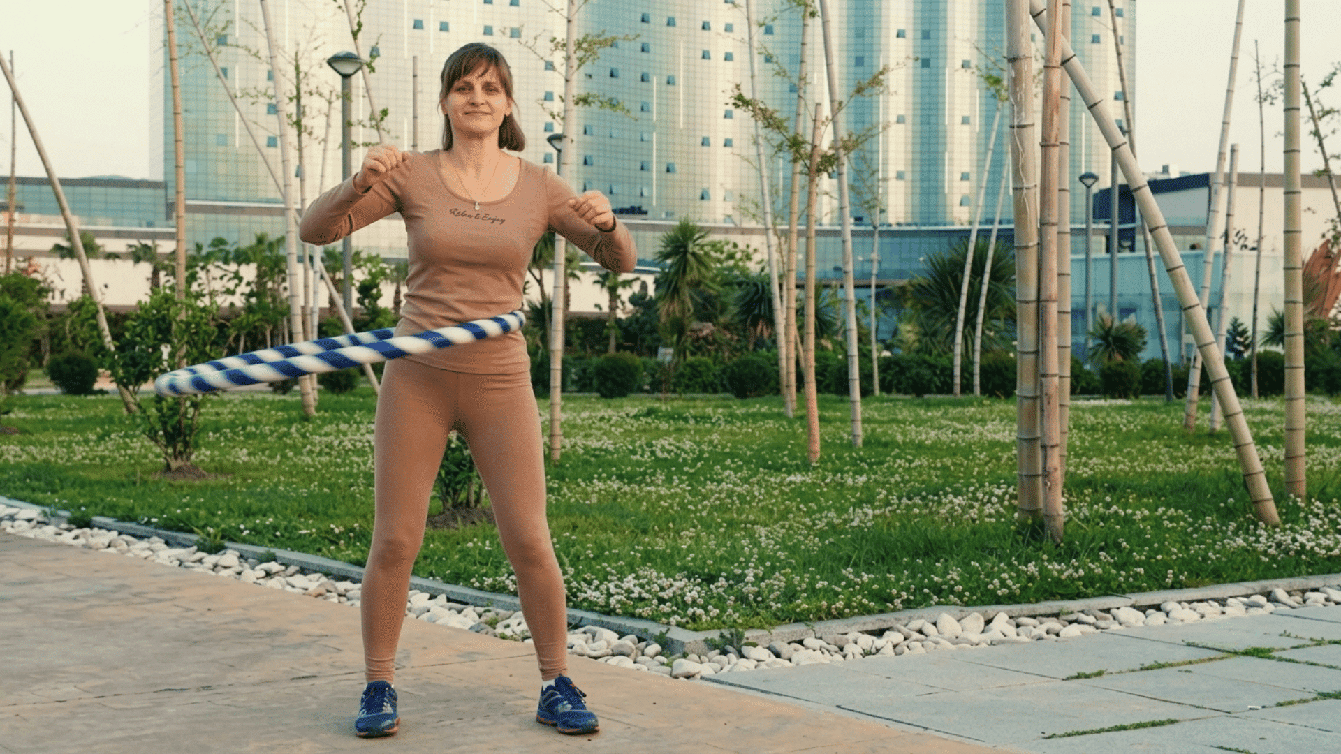 woman exercising with a hula hoop in a park surrounded by trees and modern buildings on a pleasant day