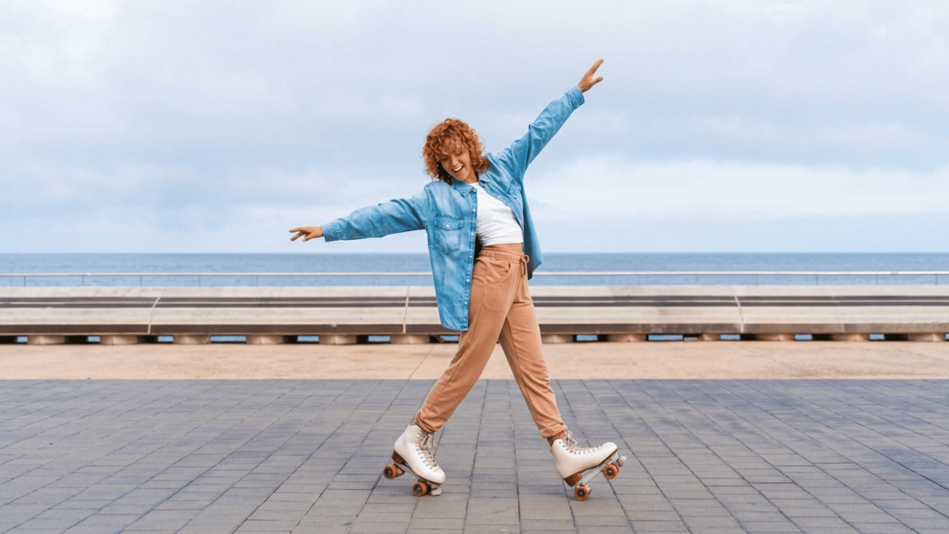 woman roller skating along a seaside promenade with arms outstretched while smiling under a cloudy sky