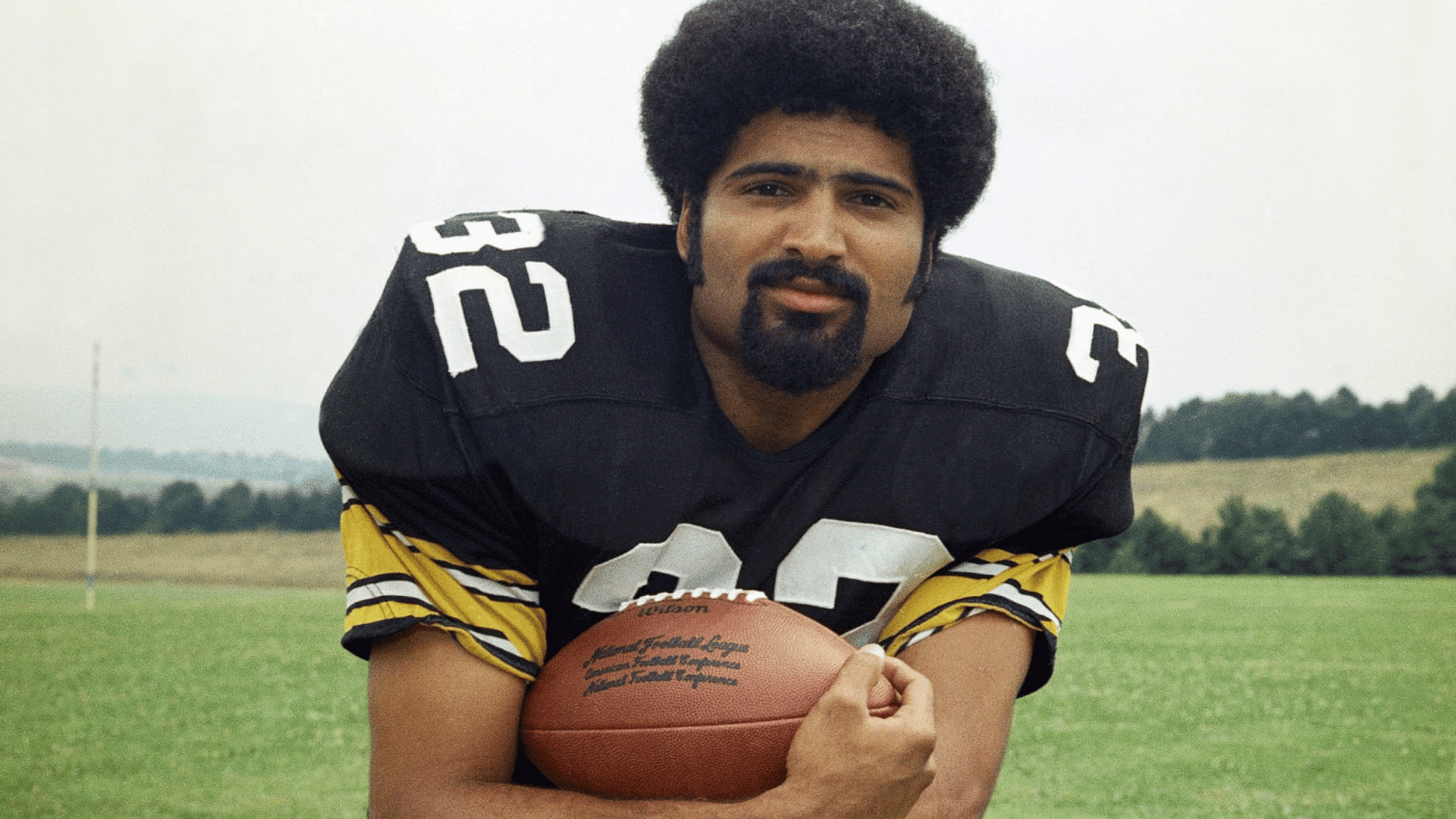 football player wearing number 32 holding a football while posing on a grassy field with trees in the distance