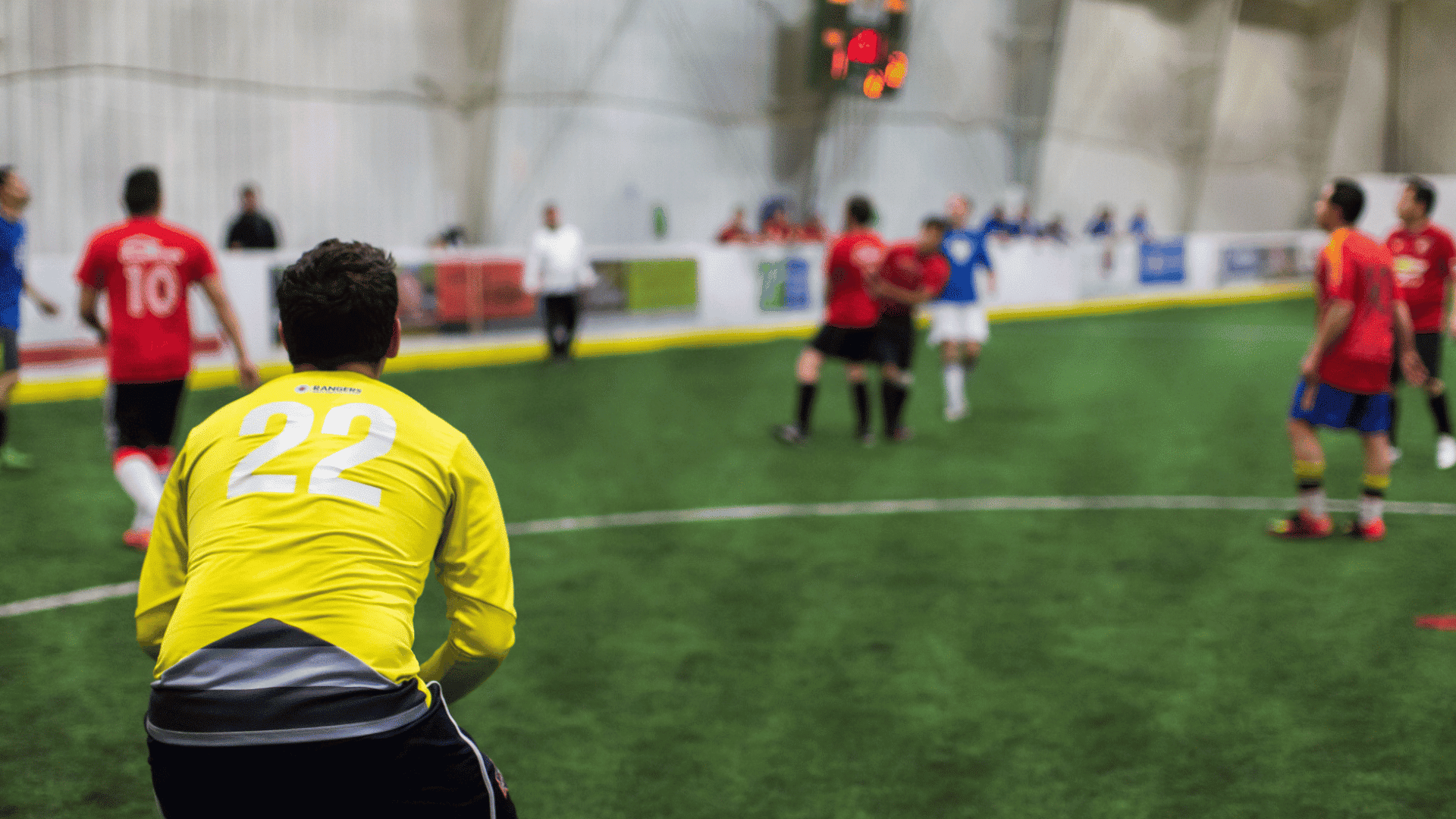 goalkeeper in a yellow jersey watching players during an indoor soccer match on a green artificial turf field