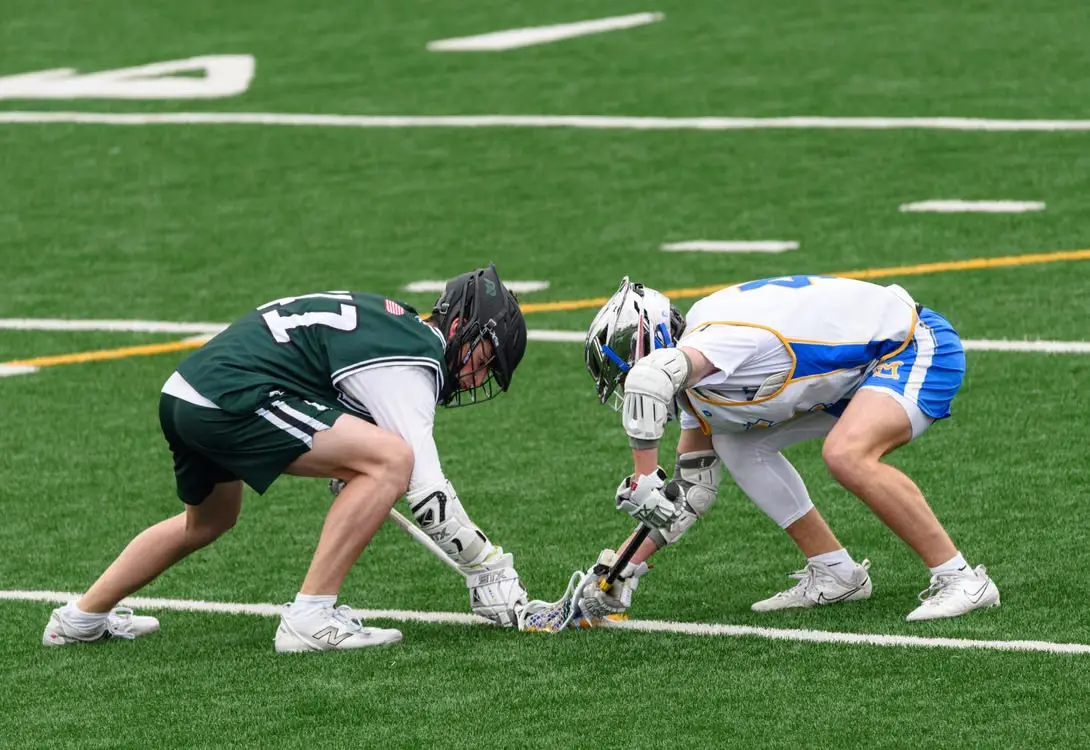Two lacrosse players facing off on green field, wearing protective gear and brightly colored uniforms