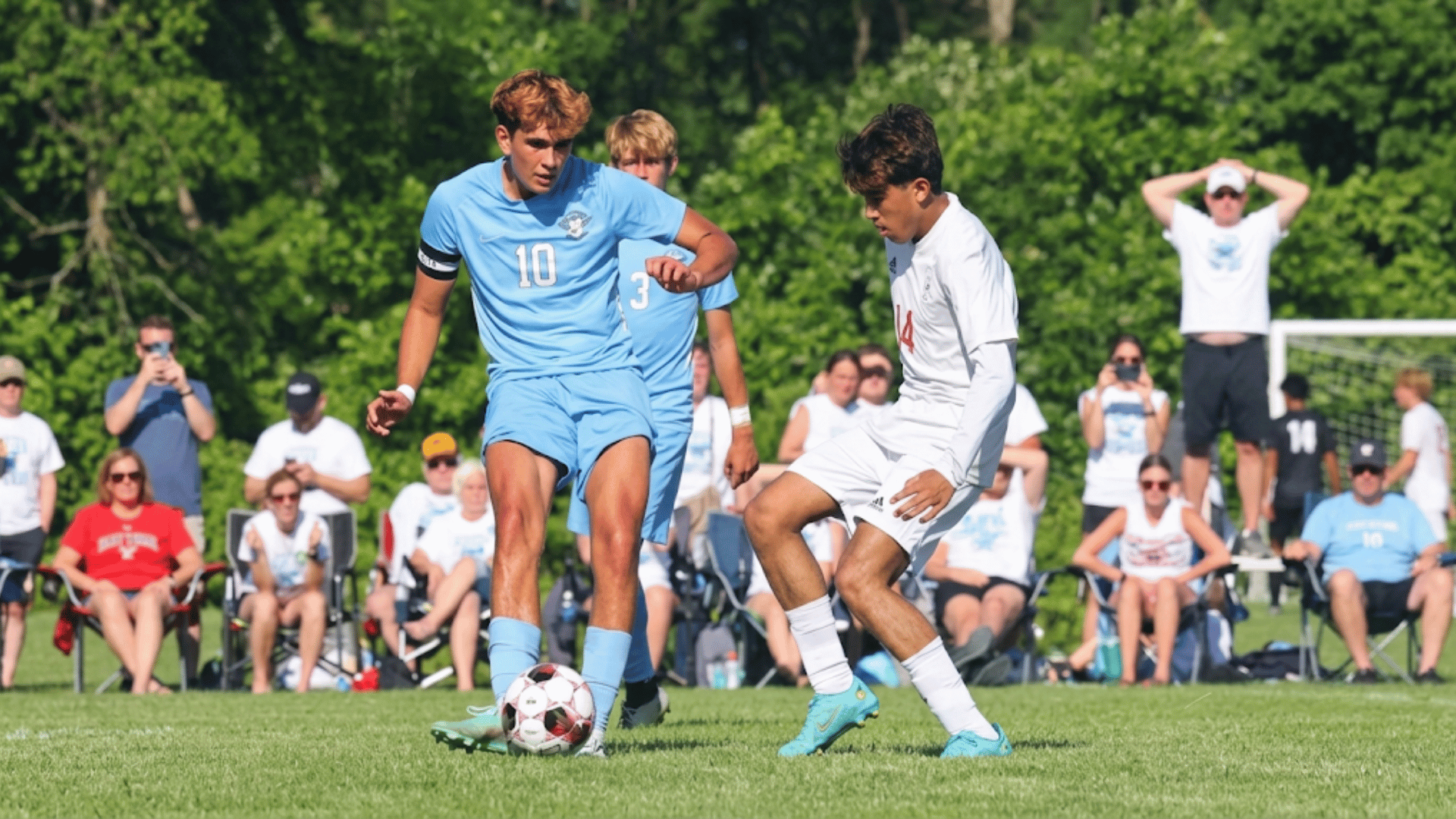 two soccer players competing for the ball on a grassy field while teammates and spectators watch from the sidelines