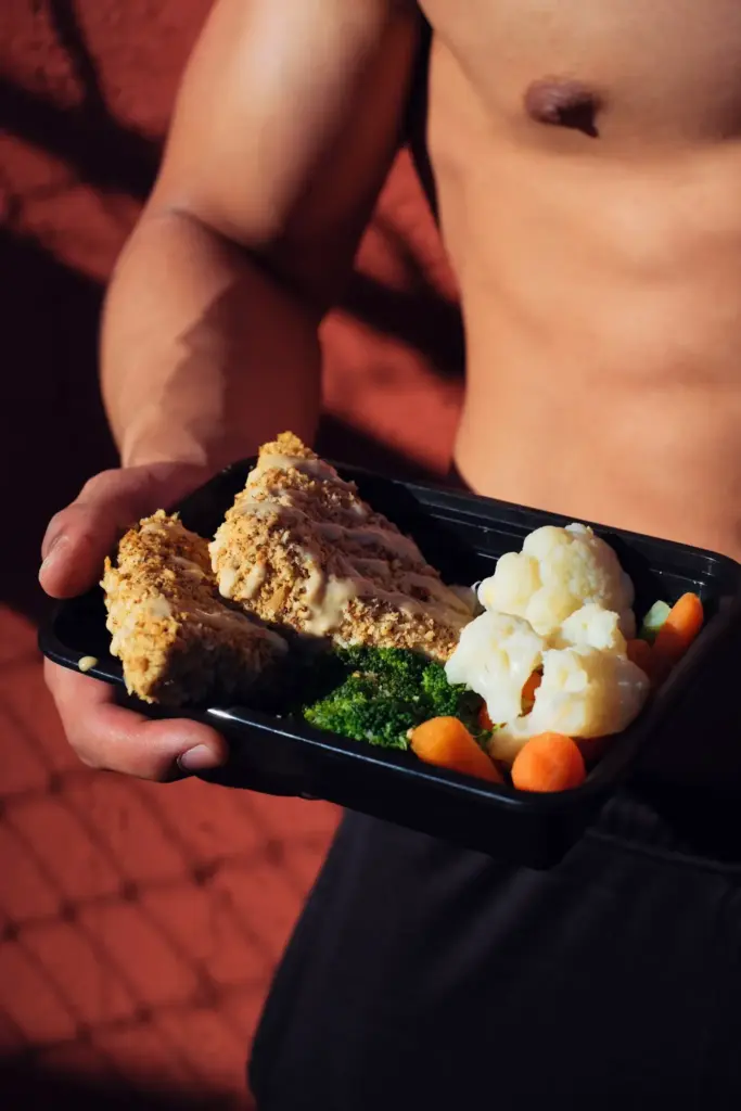 Shirtless person holding black container with breaded chicken, broccoli, cauliflower, and carrots in sunlight