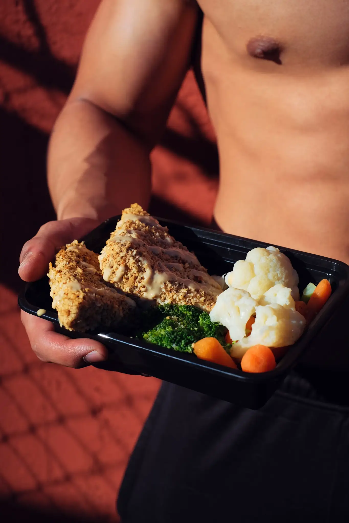 Shirtless person holding black container with breaded chicken, broccoli, cauliflower, and carrots in sunlight