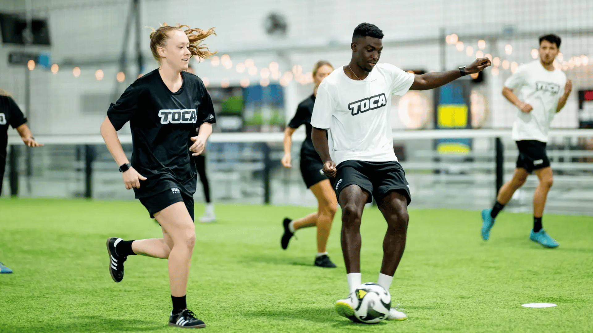 soccer players running on an indoor turf field as one player controls the ball while another follows closely