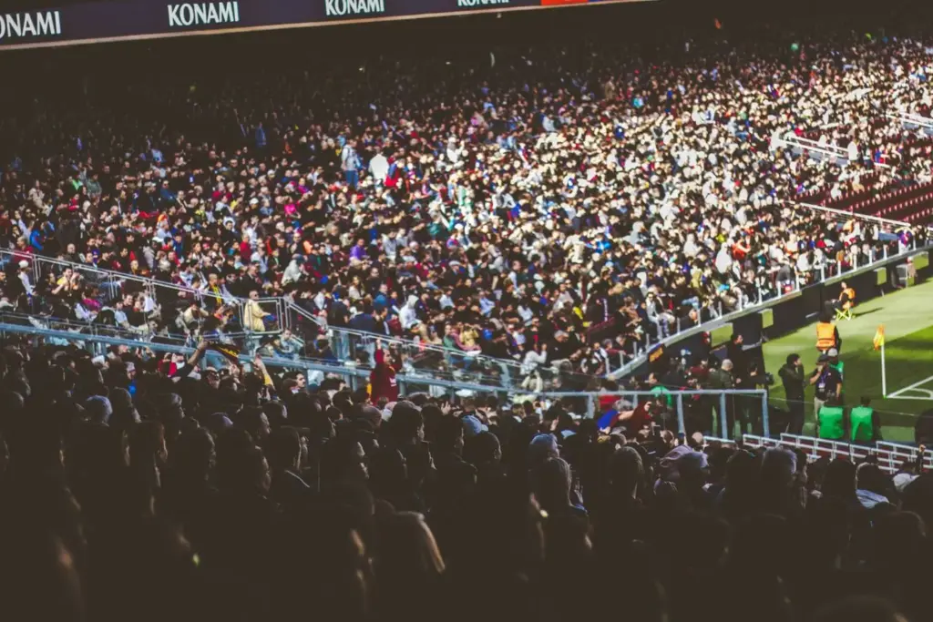Crowd of spectators seated in a stadium with green field and boundary fencing visible