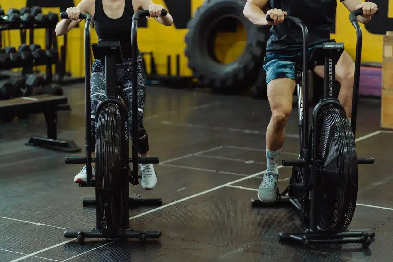 Two people exercising on air bikes in a gym with black flooring and yellow walls