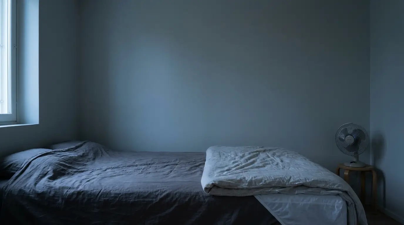 Minimalist bedroom with unmade bed, small wooden side table, and fan in dim lighting