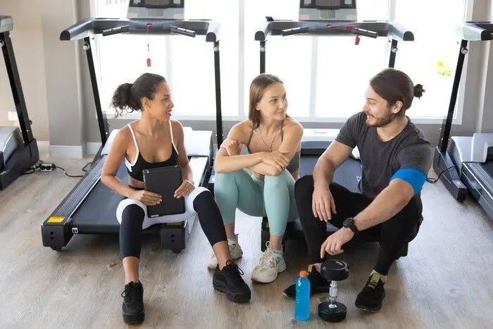 Three people sitting on gym floor talking near treadmills with water bottle and weights nearby